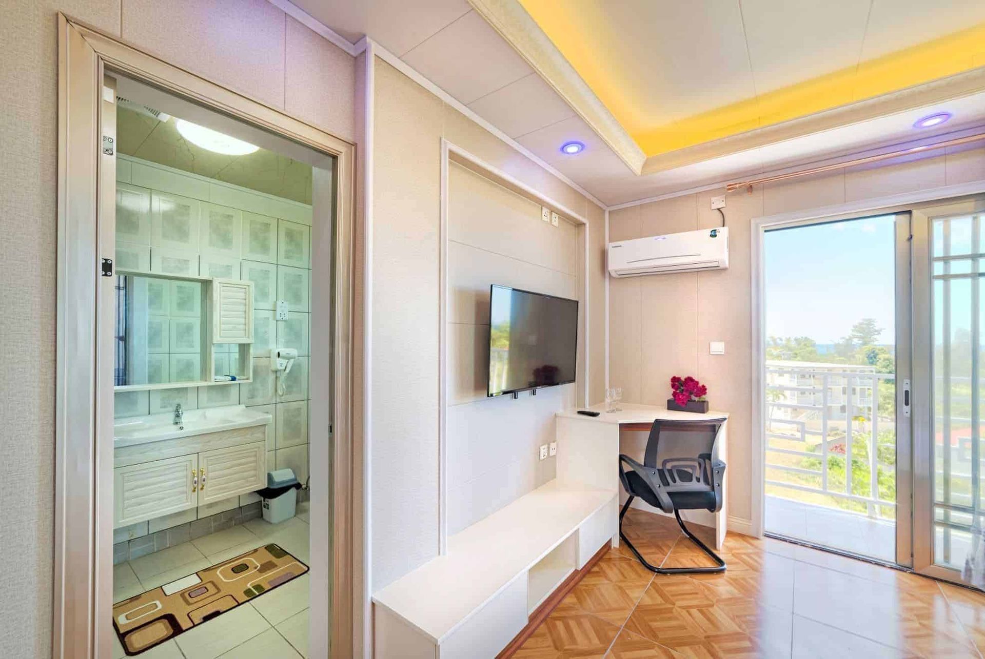 Interior view of a room with a TV, desk, and bathroom visible. Balcony with open doors.