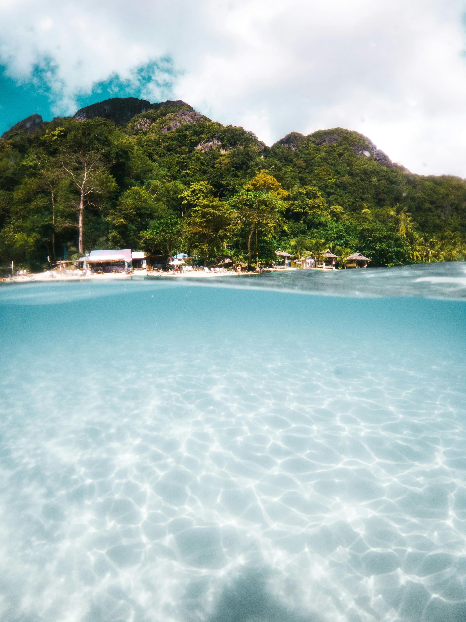Half above/below water shot of a tropical beach with clear turquoise water, white sand, and lush green mountains.