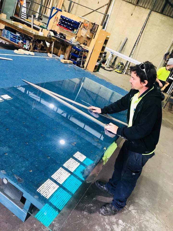 Worker measuring and cutting a large glass sheet on a blue workbench inside an industrial workshop.