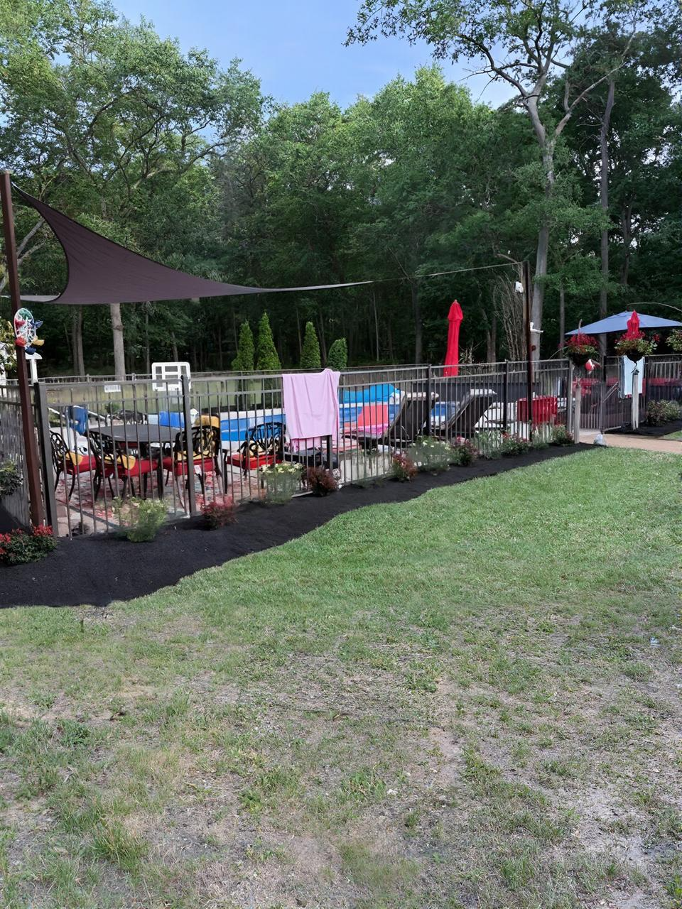 Outdoor fenced play area with colorful chairs, shade canopies, and trees beside a grassy lawn