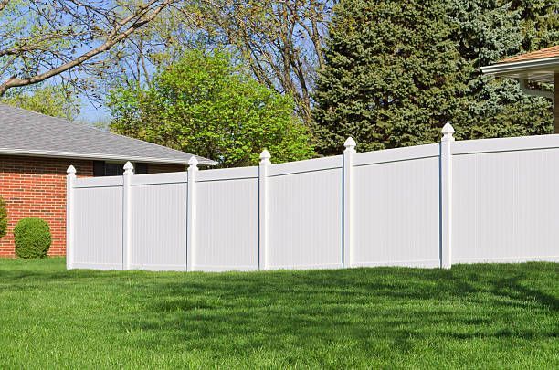 A white vinyl privacy fence with pointed post caps stands in a green yard next to a brick house