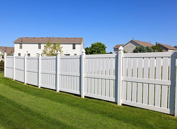 A white vinyl privacy fence stands on a mowed green lawn in a suburban neighborhood under a clear blue sky