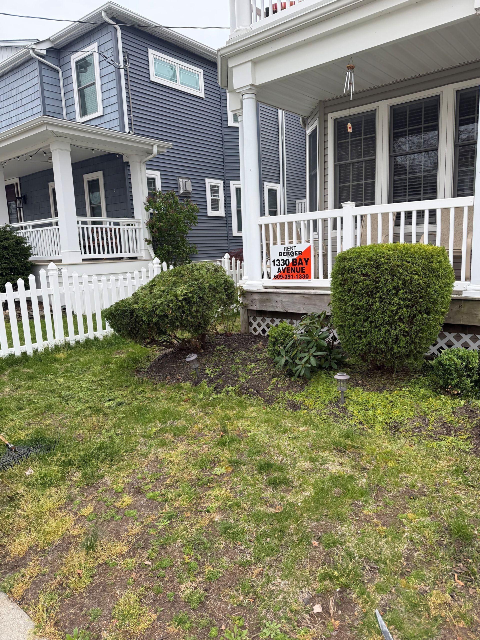 Front porch of a gray house with white trim, a white picket fence, and small shrubs in front