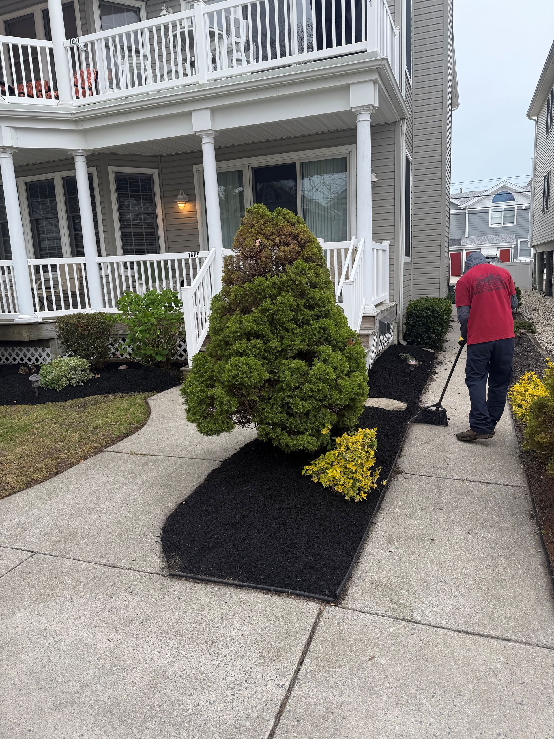 Sidewalk beside gray house with white porch and trimmed shrubs, a person in a red jacket walking away