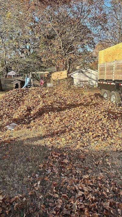 A large pile of dry brown autumn leaves sits on a lawn next to a truck, with trees and a white house in the background