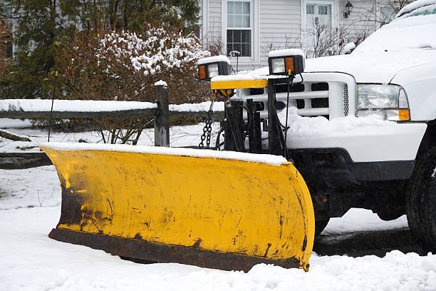 A white truck with a yellow snow plow attachment parked on a snow-covered ground in front of a house
