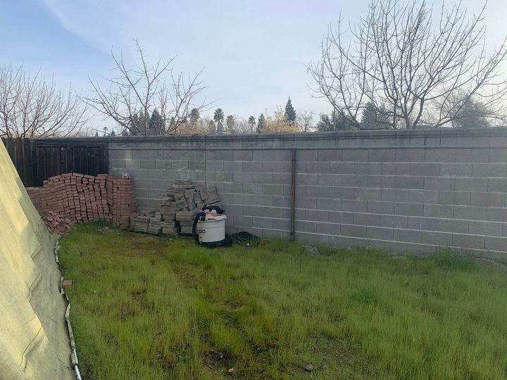 A grassy yard features a gray concrete wall, a stack of bricks, and a shop vacuum sitting on the grass.