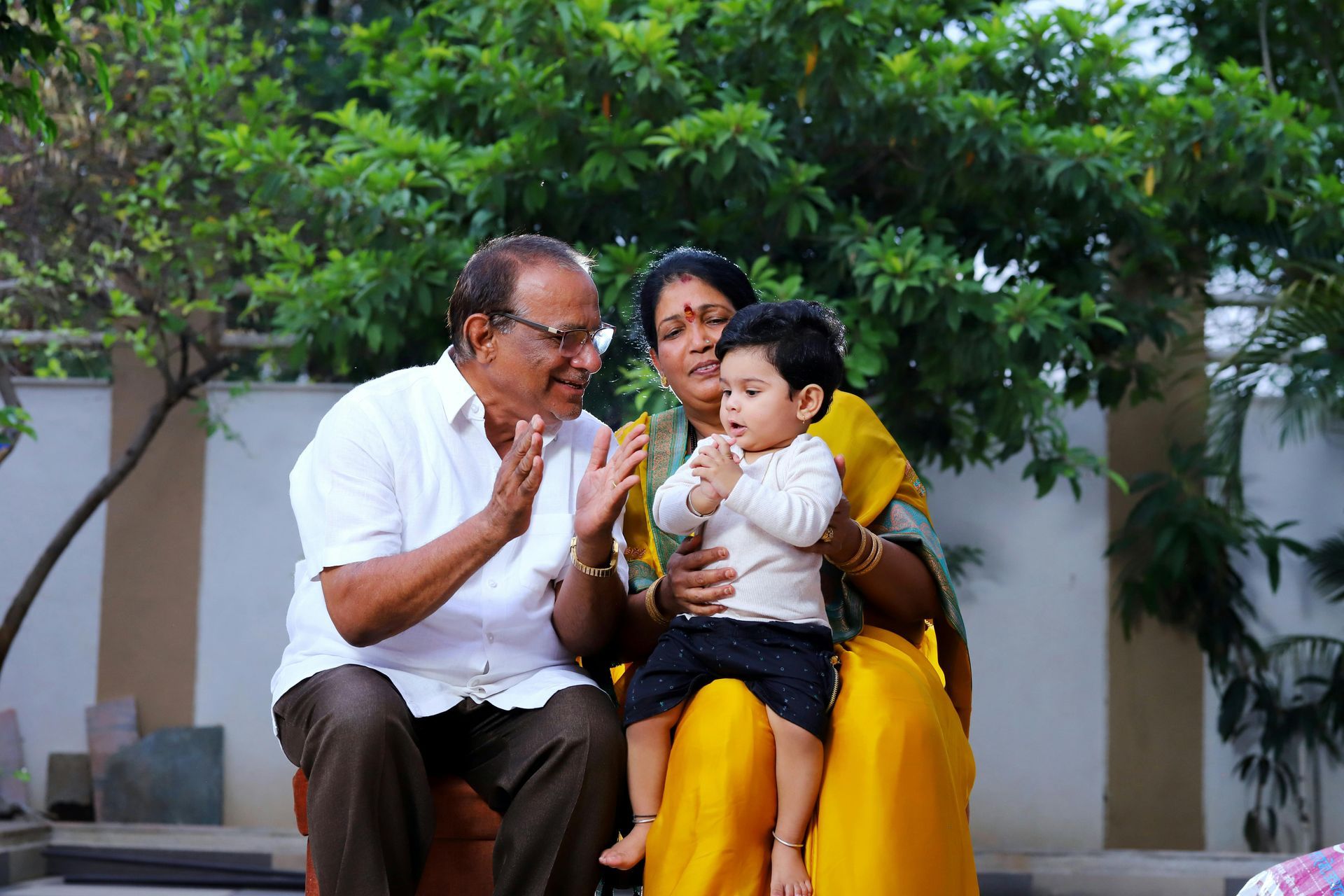 Three people sitting outdoors, smiling and sharing a moment on a bench near green trees.