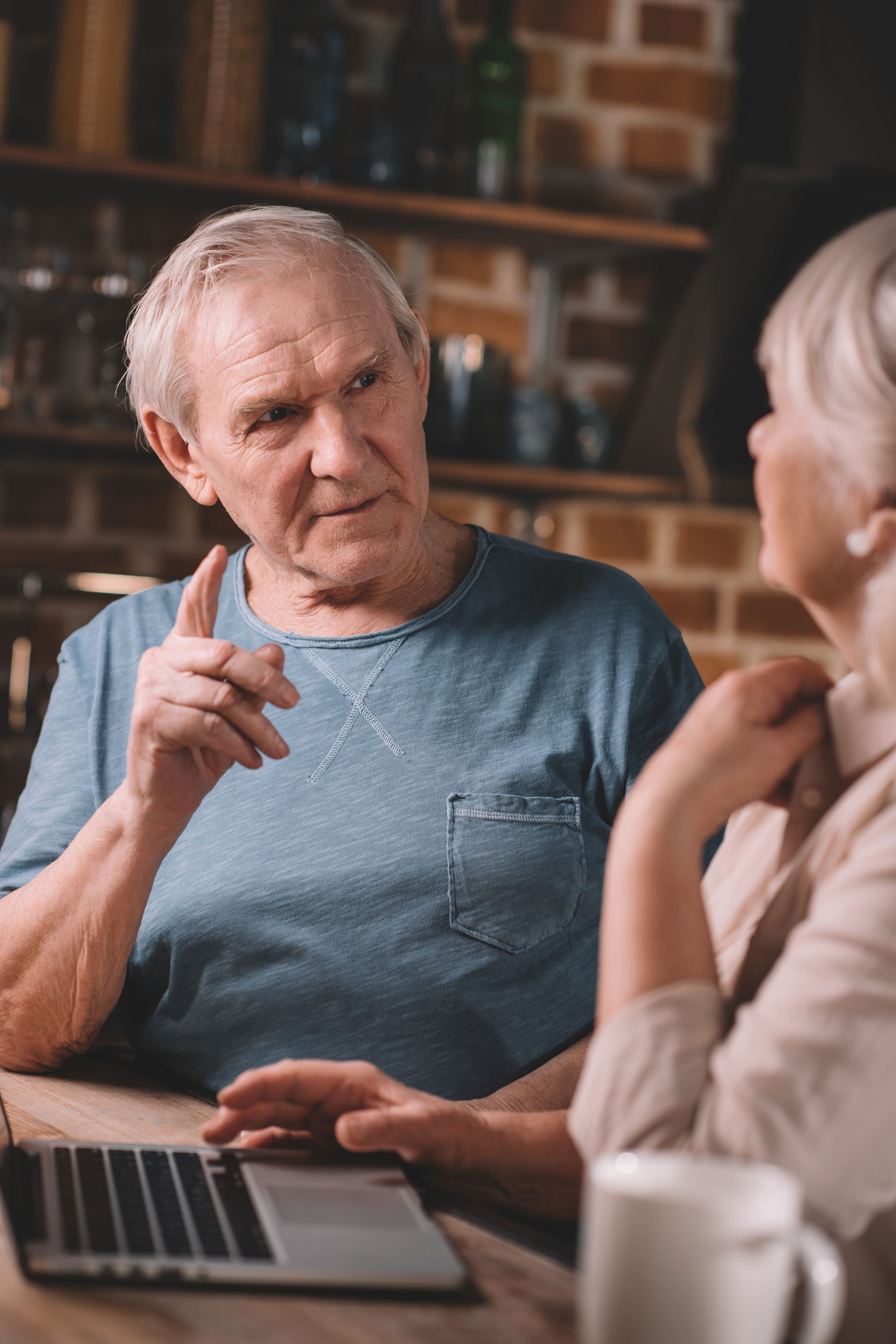 Two people talking at a table with a laptop and coffee mugs in a brick-walled café