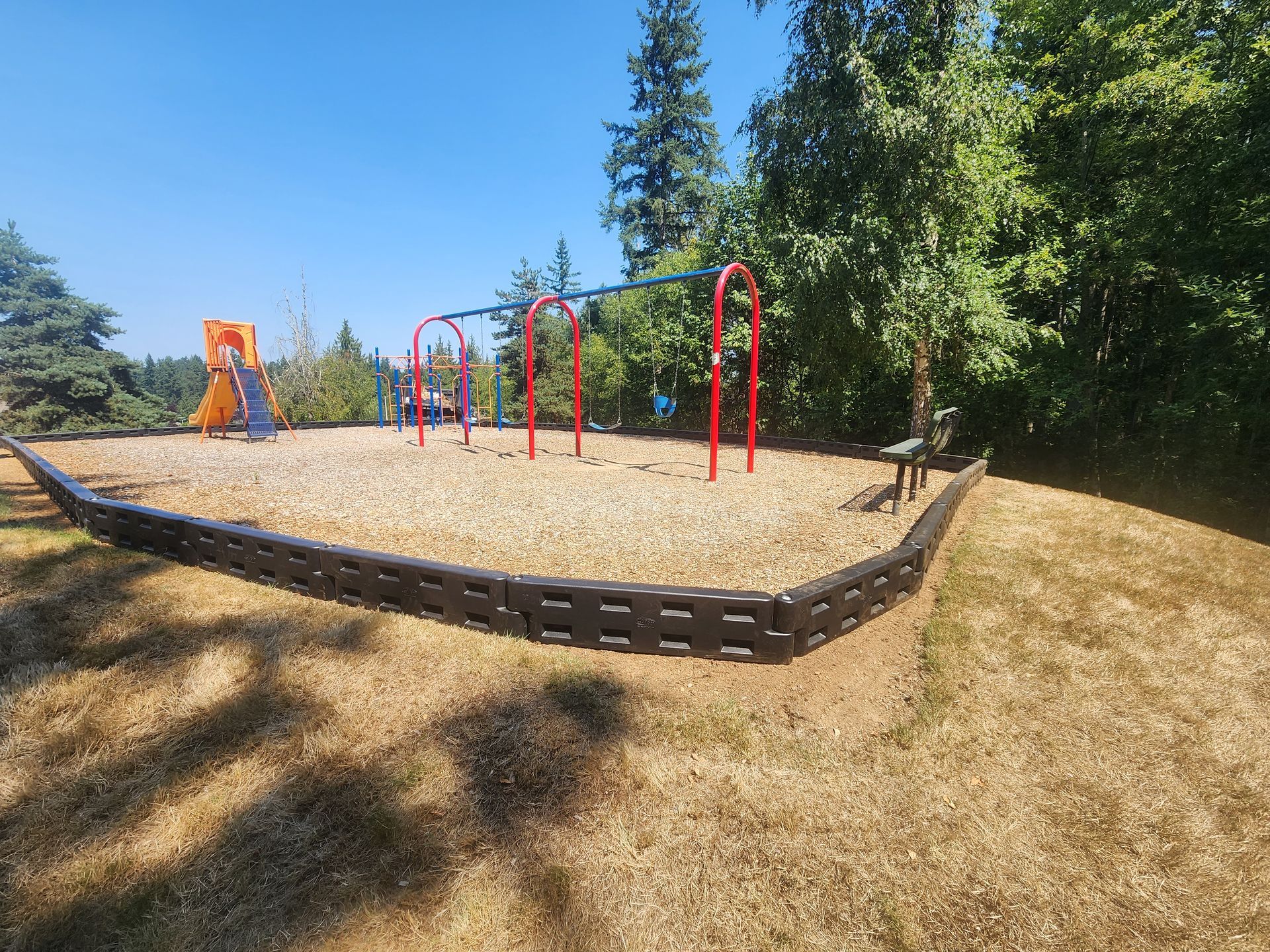 Playground with swings and slide, surrounded by wood chips and black border, under a blue sky.