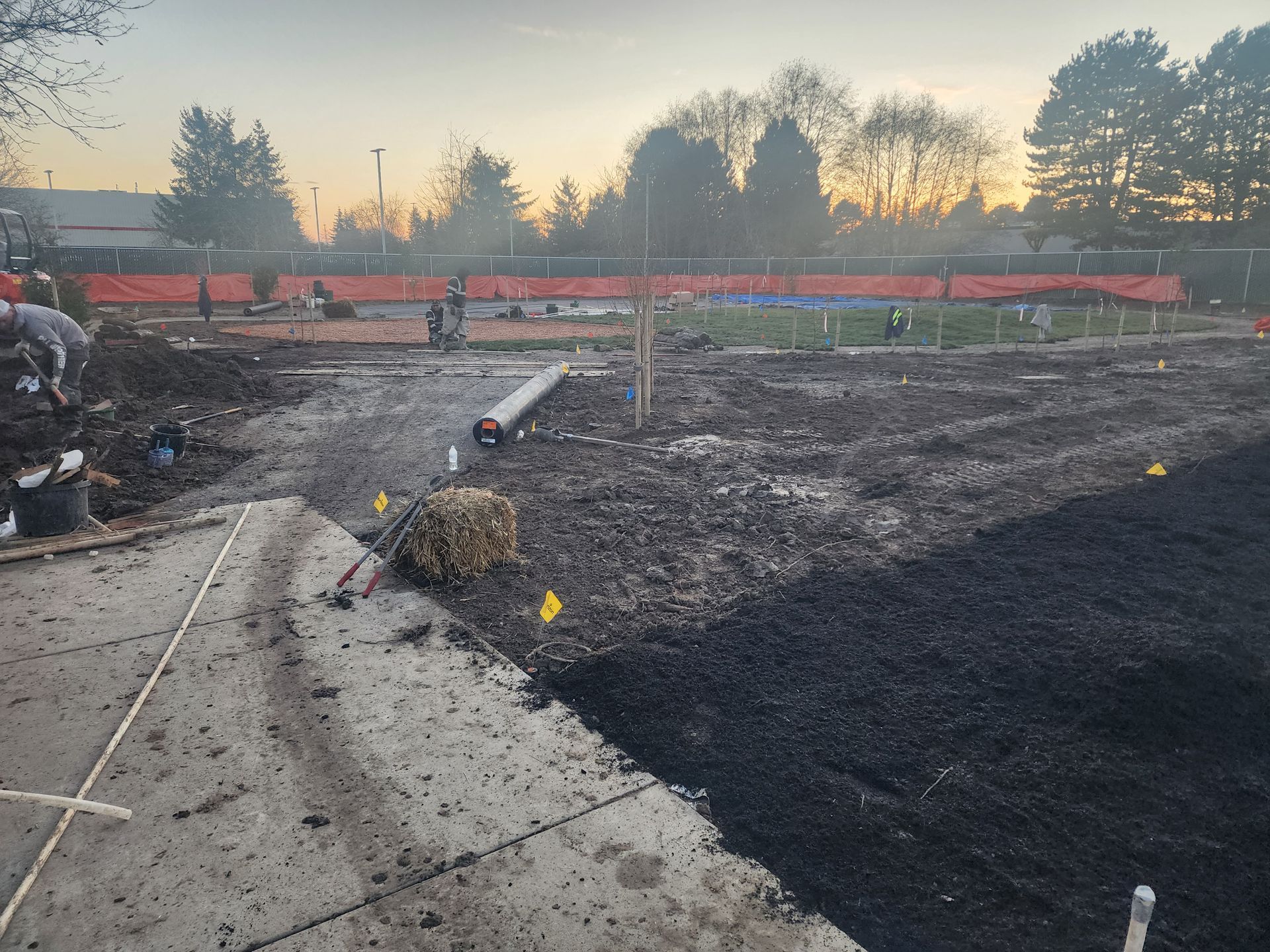 Construction site with dirt, pipes, and orange fencing; a partially paved area in the foreground.
