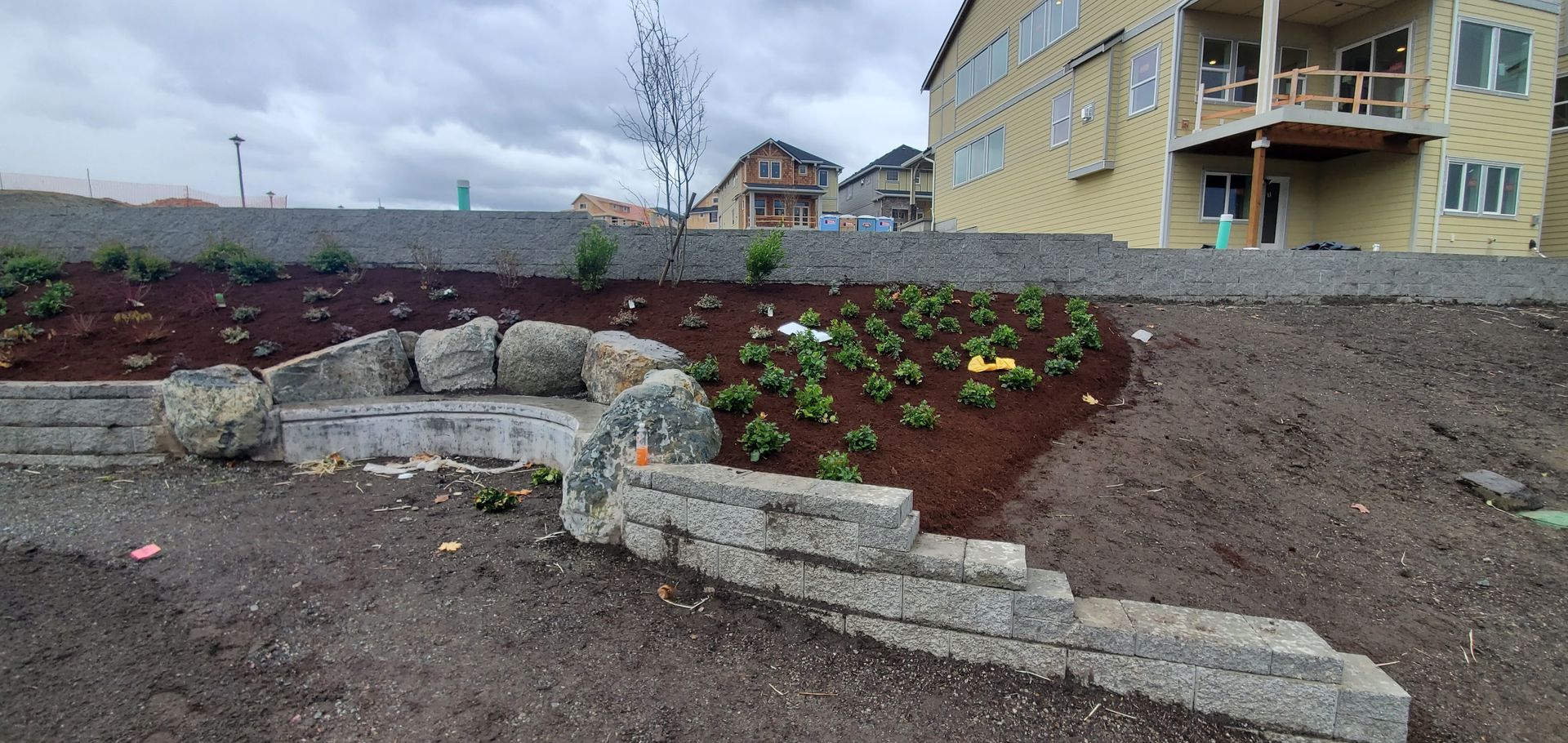 Landscaped hillside with stone retaining walls, plants, and a residential building under a cloudy sky.
