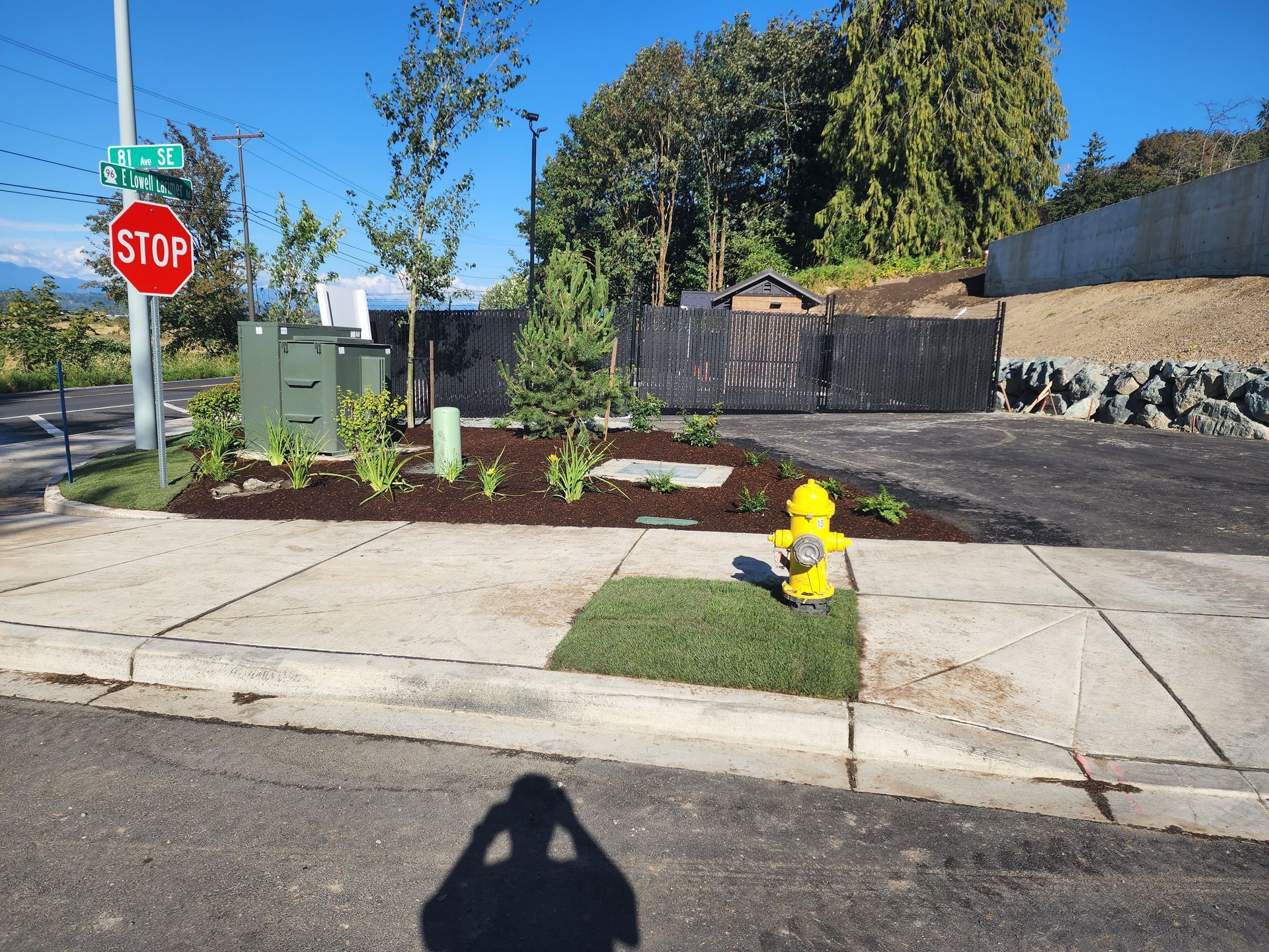Street corner with a stop sign, utility boxes, landscaping, a fire hydrant, and a partially paved driveway.