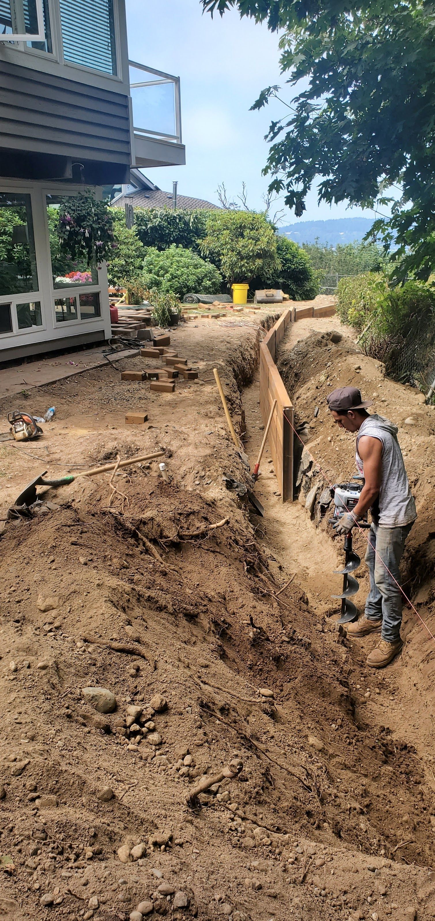 Man using a jackhammer in a trench next to a wooden barrier. Construction site with a house.