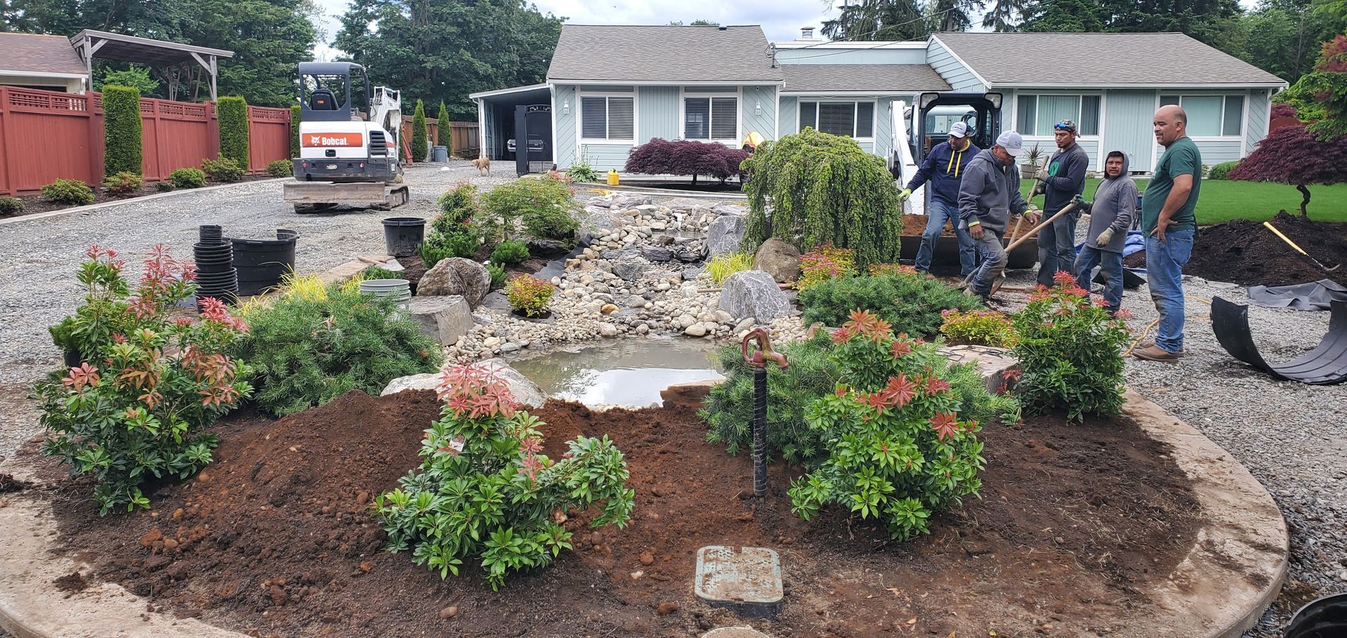 A small crew landscaping around a small pond and bushes in front of a house.