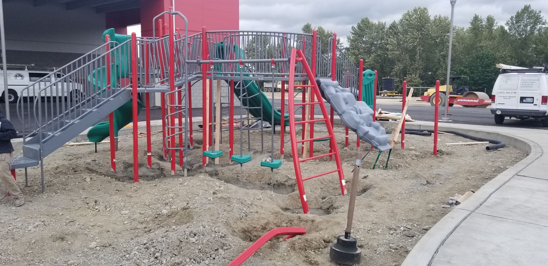 A playground with red and silver equipment surrounded by gravel, next to a paved area.