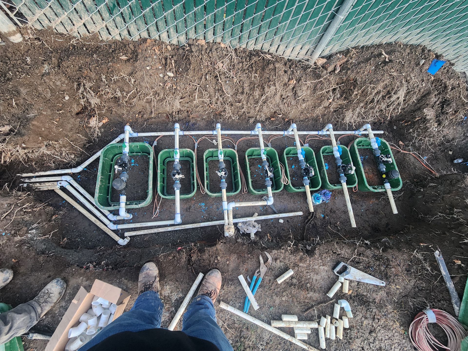 Irrigation system components laid out in a trench, including valves and piping, near a fence, with tools nearby.