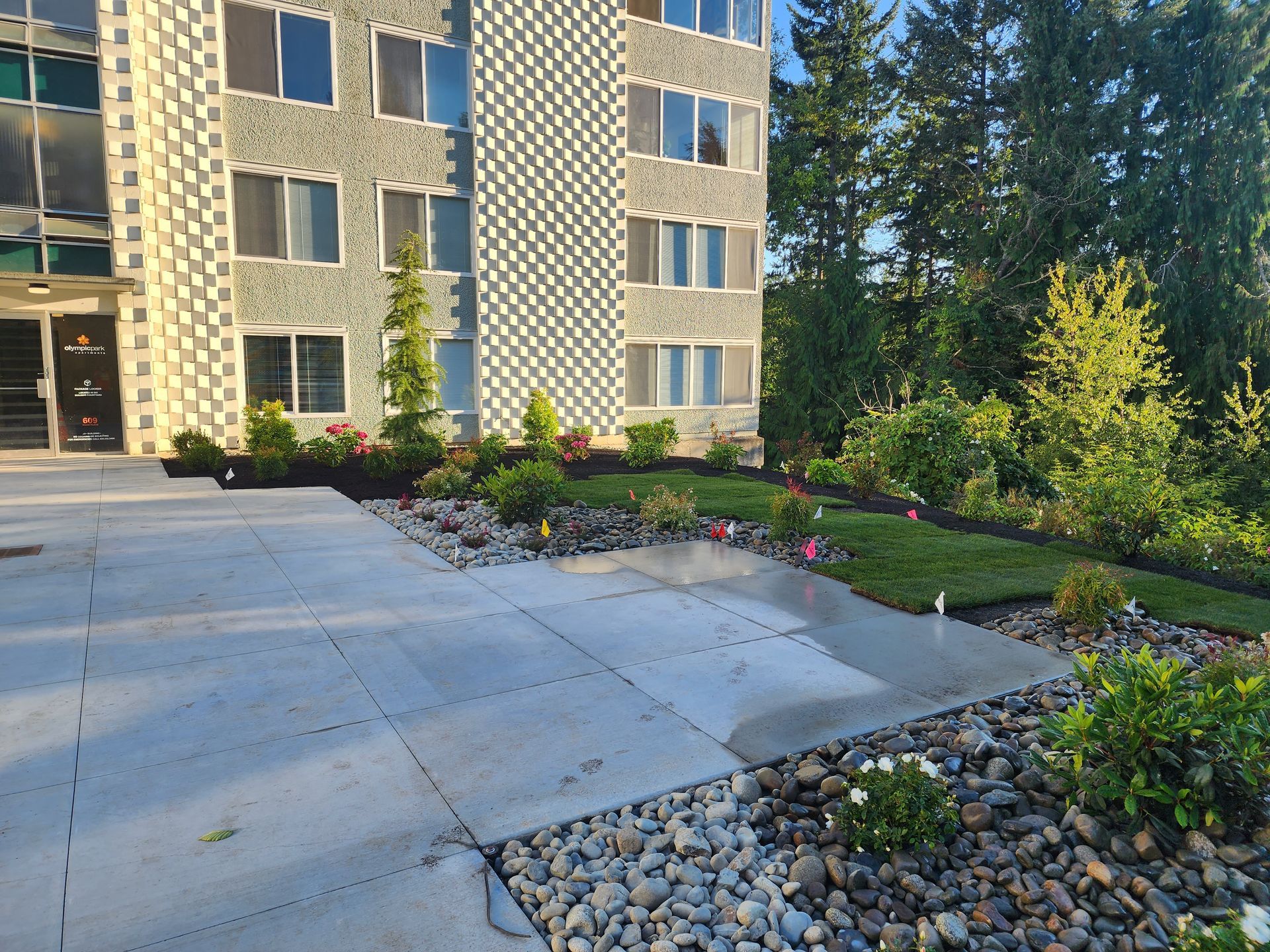 Apartment building with landscaping, including rocks, flowers, and greenery. Sunlight illuminates the scene.