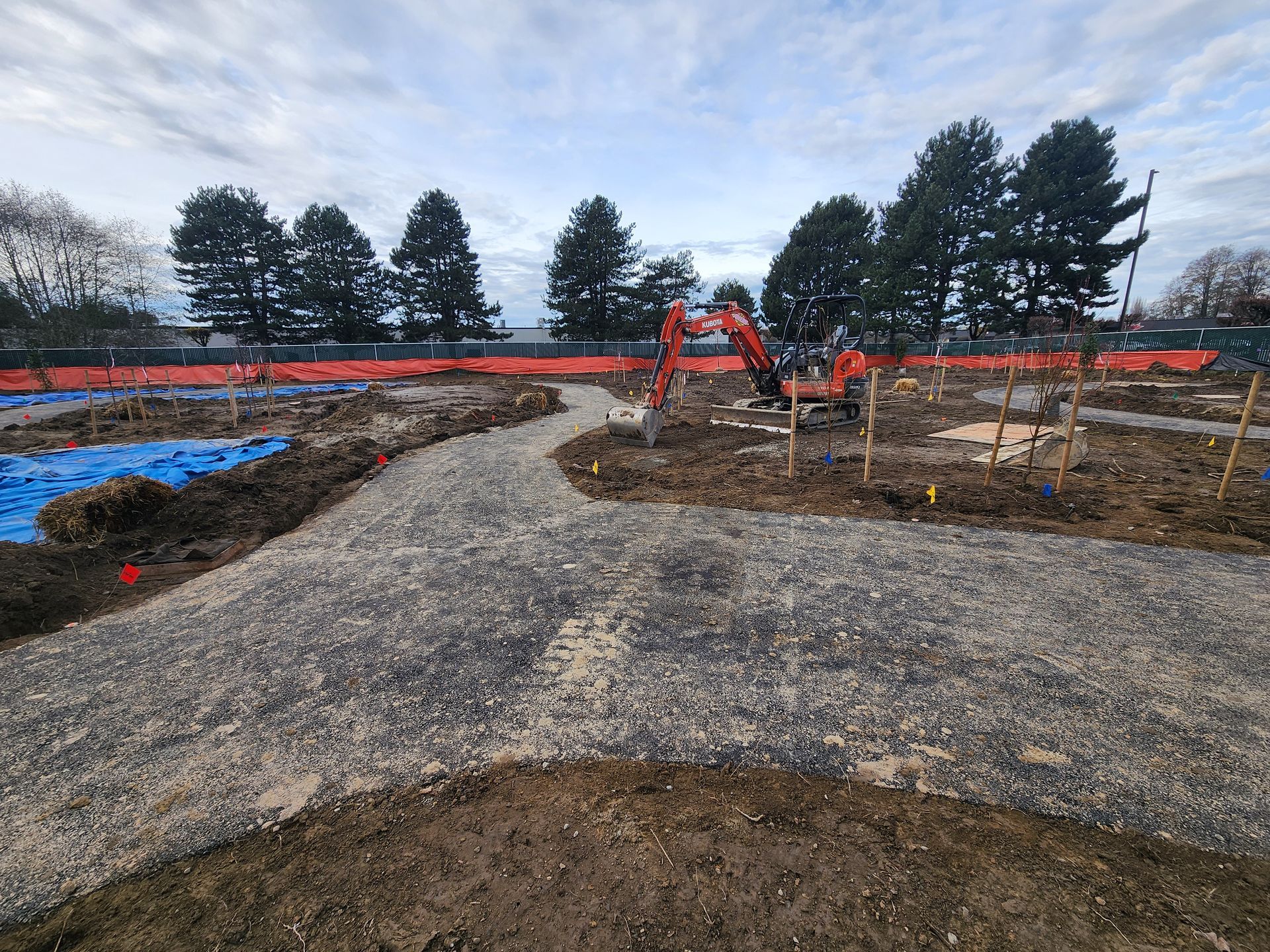 A gravel path curves through a construction site, an excavator in the distance, blue tarp, orange fencing.