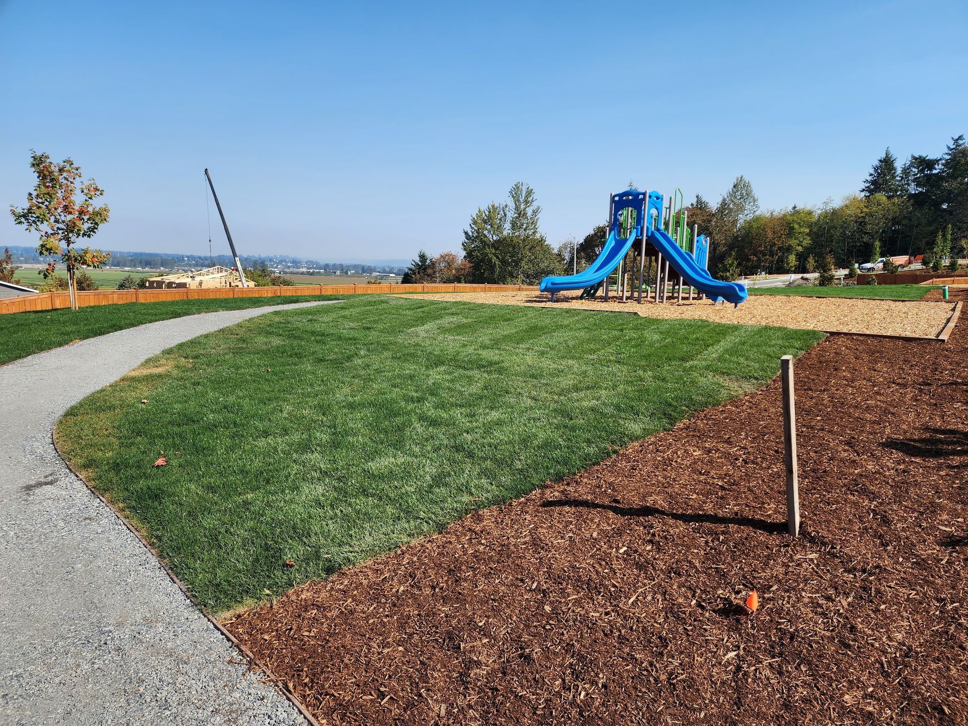A blue playground slide on a grassy hill next to a wood-chip ground covering and a gravel path, under a clear sky.