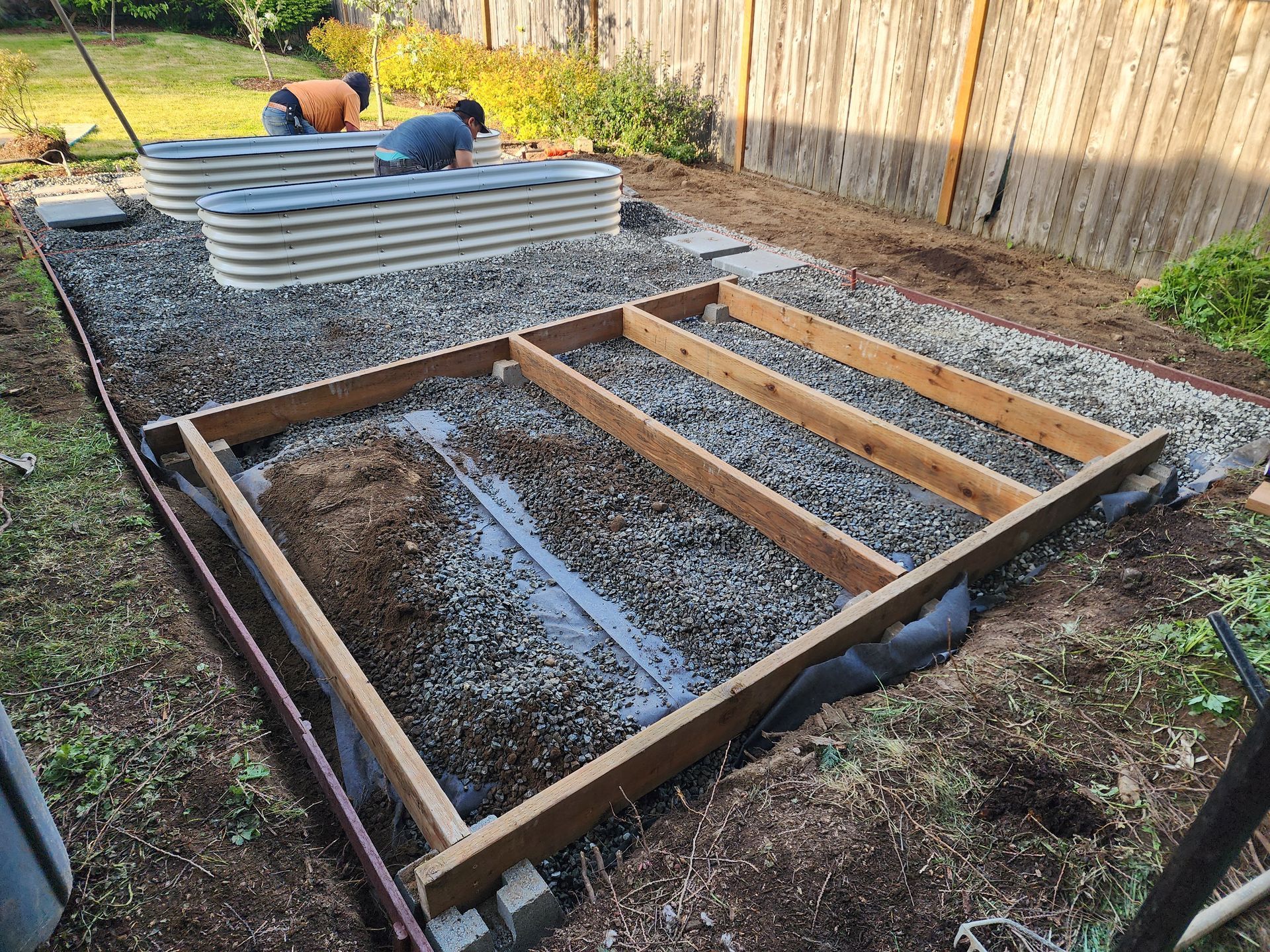 Two people installing raised garden beds. A wooden frame sits on gravel.