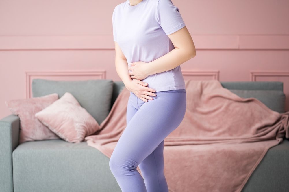 Woman in lavender activewear holding her stomach, appearing to be in pain, by a pink couch — Barolin Physiotherapy Services in Bundaberg South, QLD