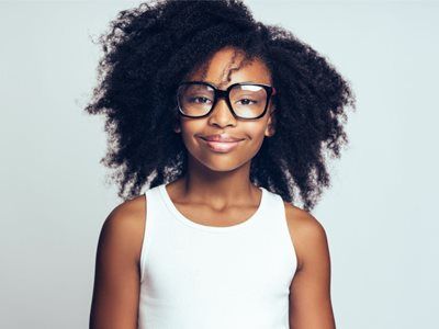 Girl with curly hair wearing glasses smiles at the camera, white background.