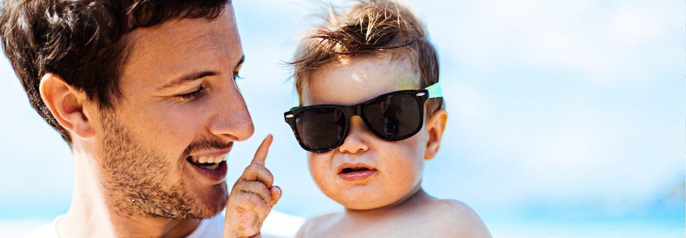 Man pointing at a baby wearing sunglasses on a beach; baby is squinting with a serious expression.