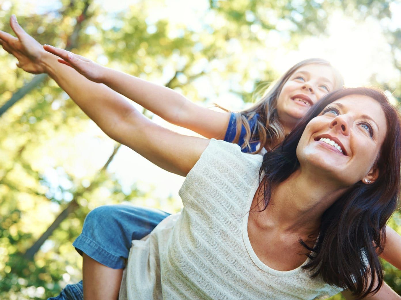 Woman carries a smiling child on her back, arms outstretched, outdoors.