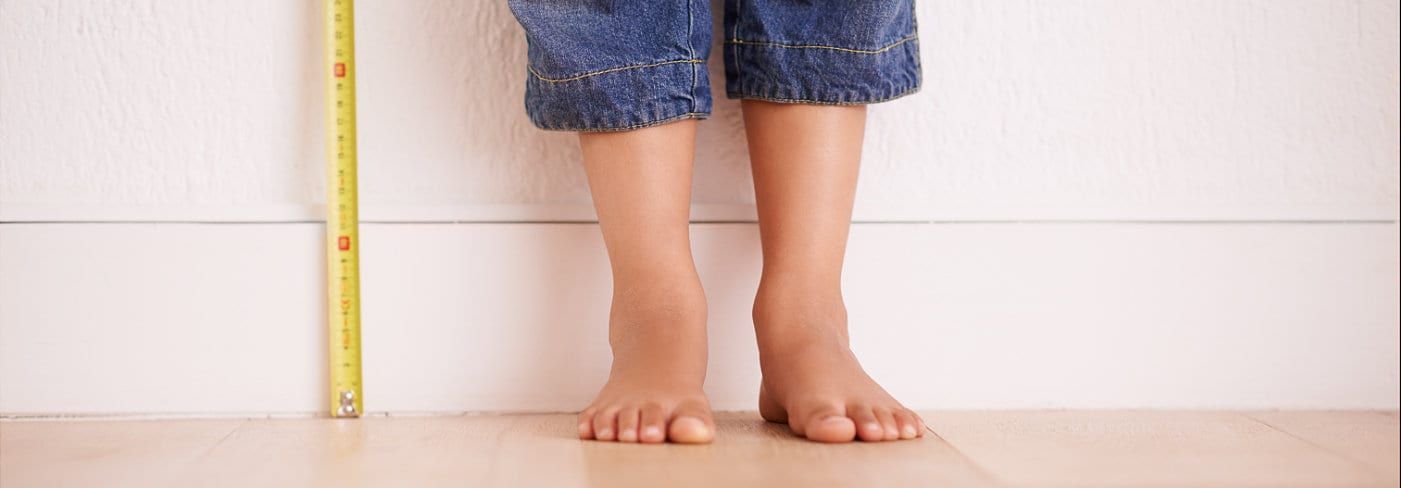 Child's bare feet and blue jean shorts, standing in front of a white wall, with a measuring tape on the side.