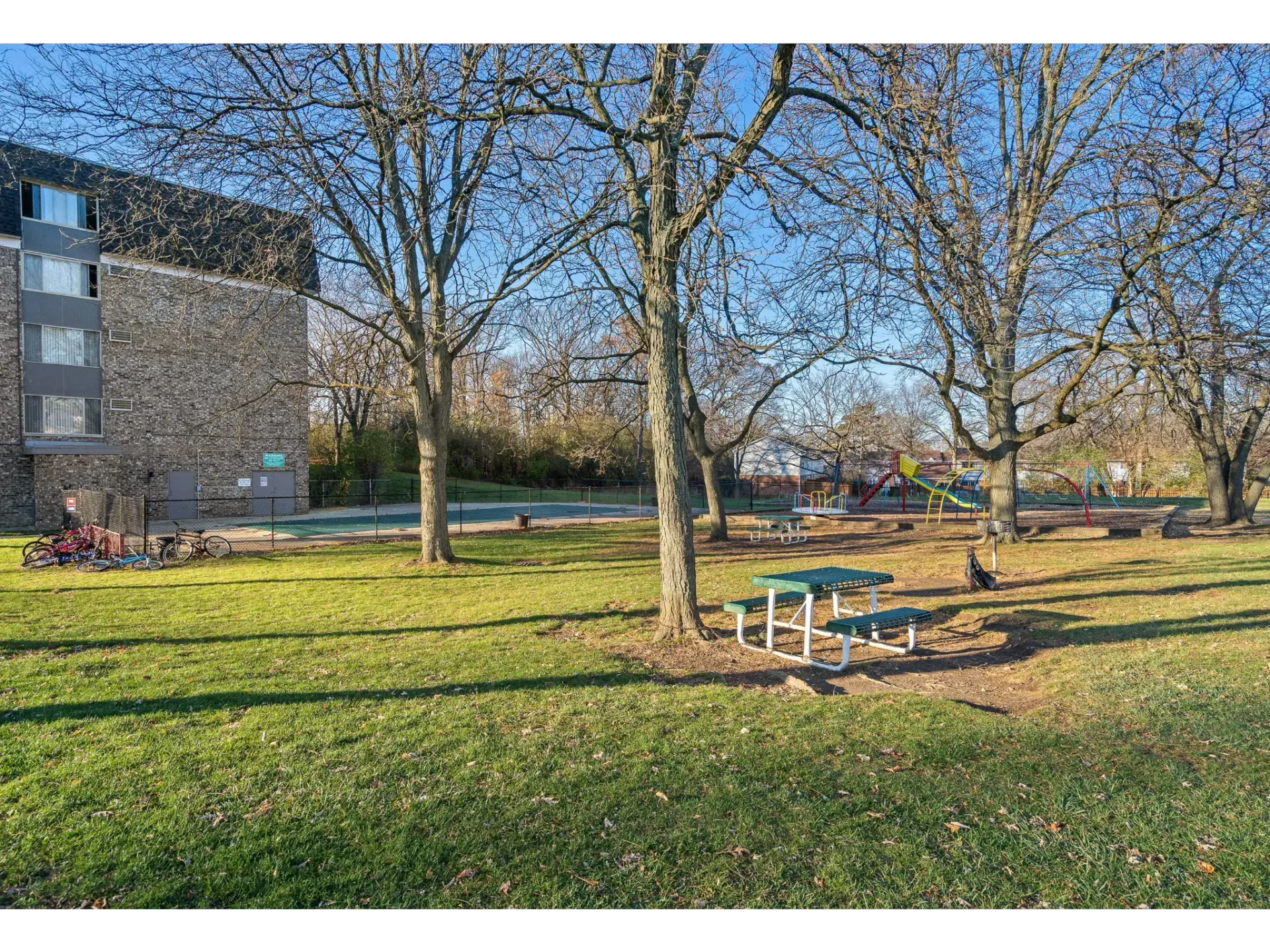 View of a community park with trees, picnic tables, playground, and a building in the background at Bartlett Lakes, offer apartments for rent in Bartlett, IL.