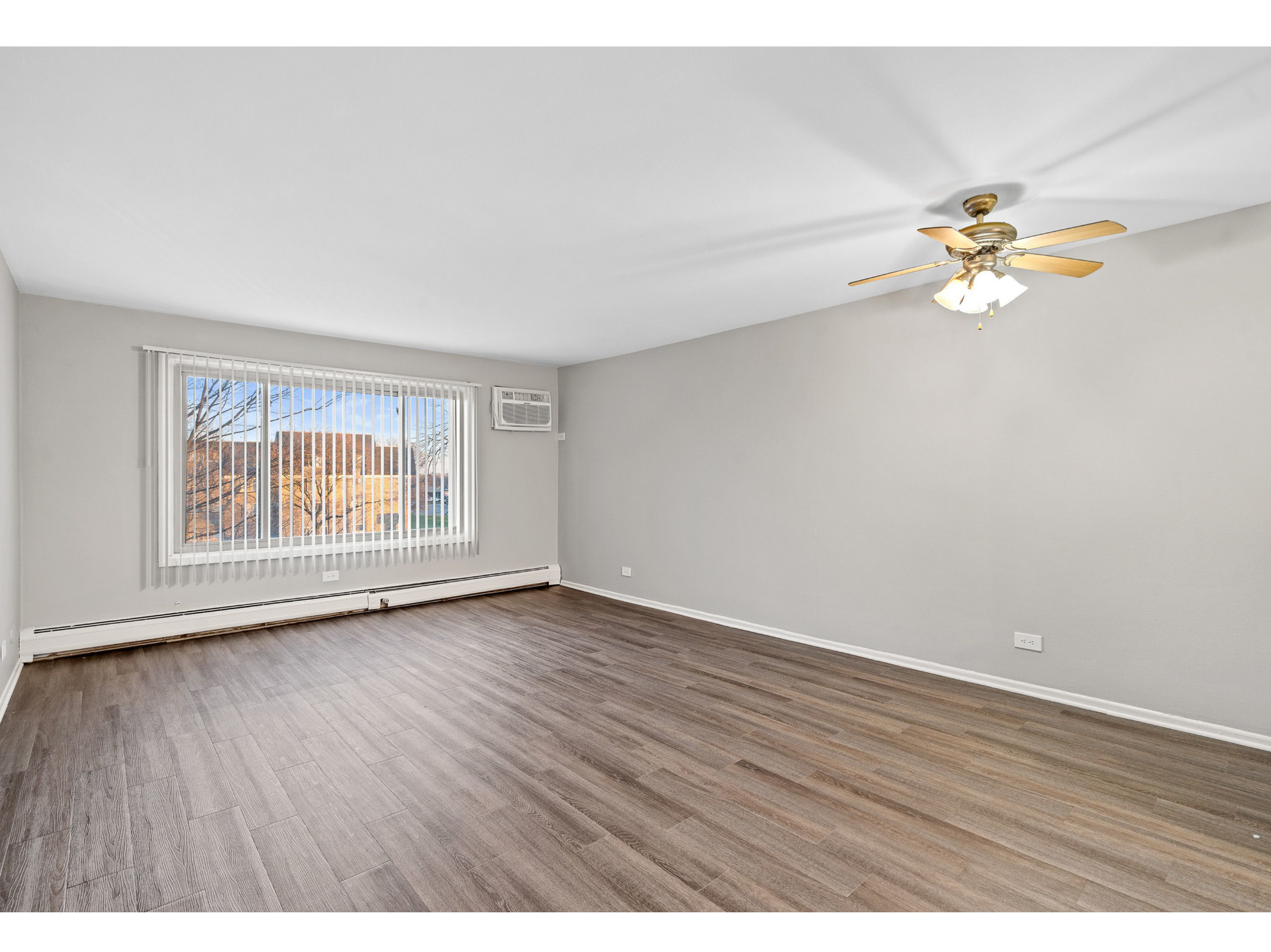 Empty living room with large window, vertical blinds, ceiling fan, and wood-look flooring.