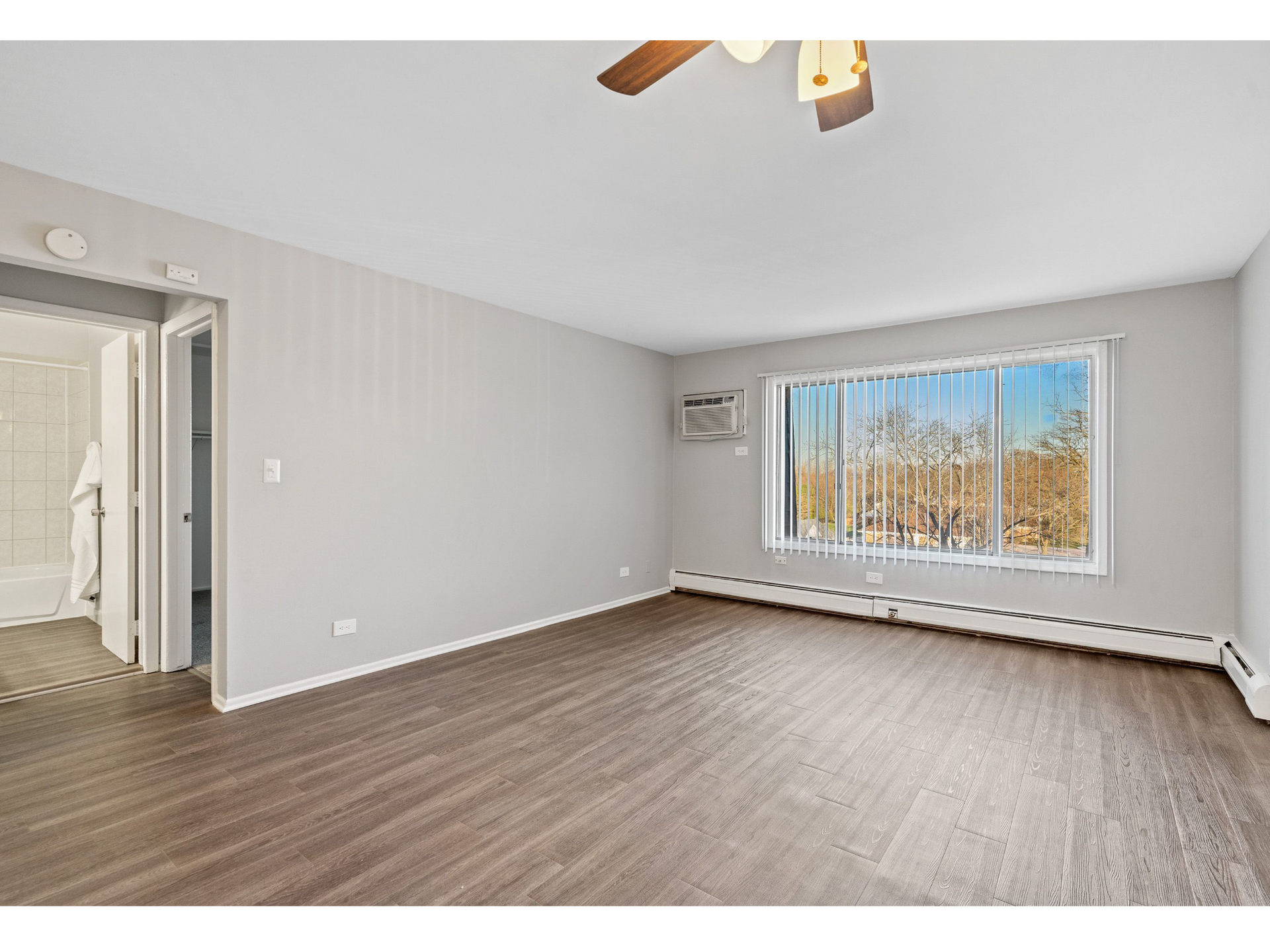 Empty living room with wood-look flooring and a large window with vertical blinds.