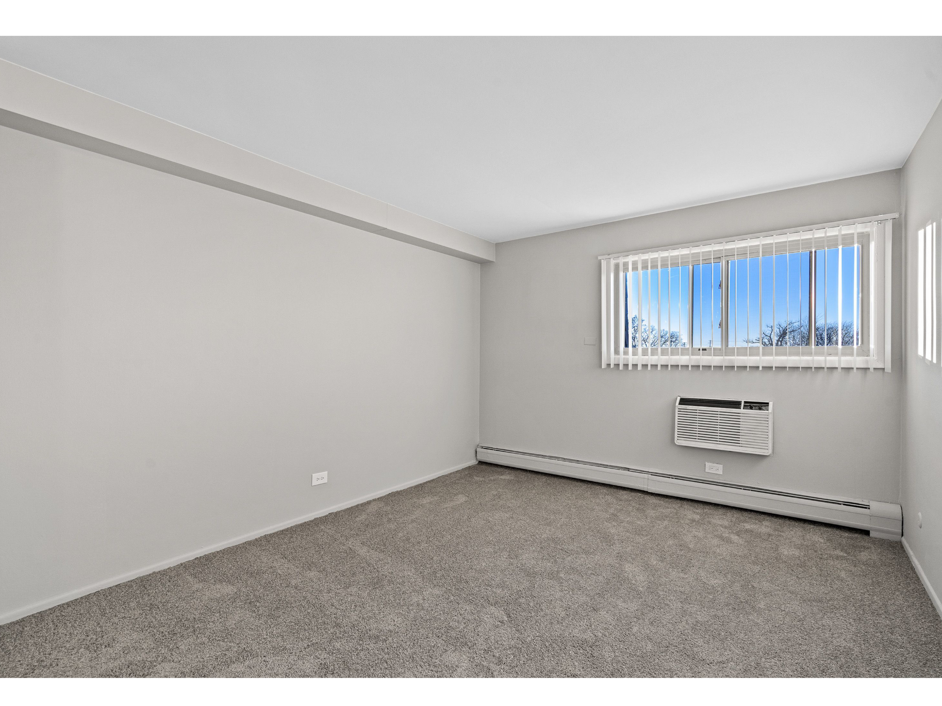 Empty apartment room with window, vertical blinds, and wall-mounted AC unit.