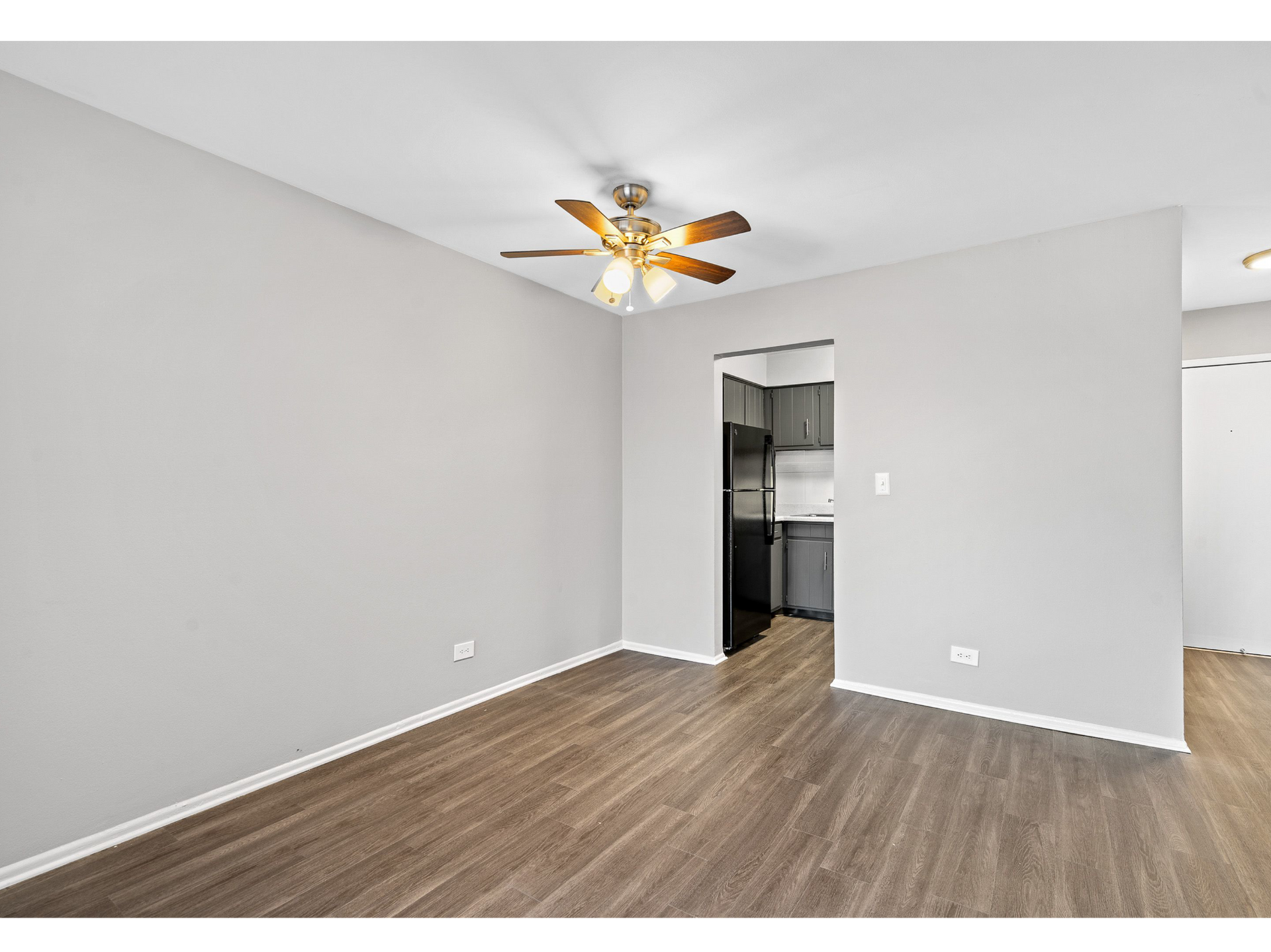 Empty living room with gray walls, wood-look flooring, ceiling fan; kitchen entrance visible.