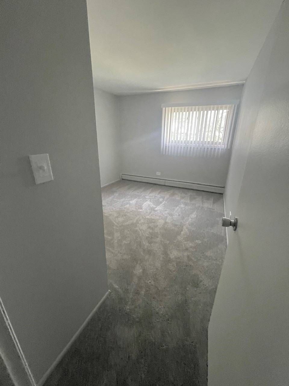 Empty bedroom with gray walls, carpet, baseboard heater, and a window with vertical blinds.