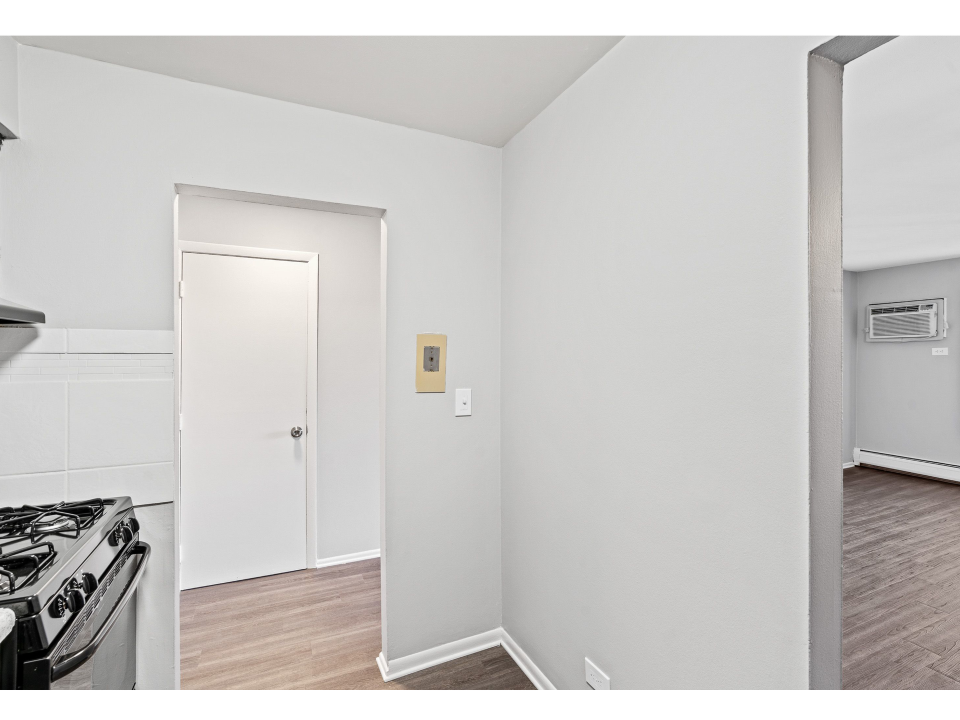 Kitchen area with a stove on the left, a white door in the center, and a view into the living area.