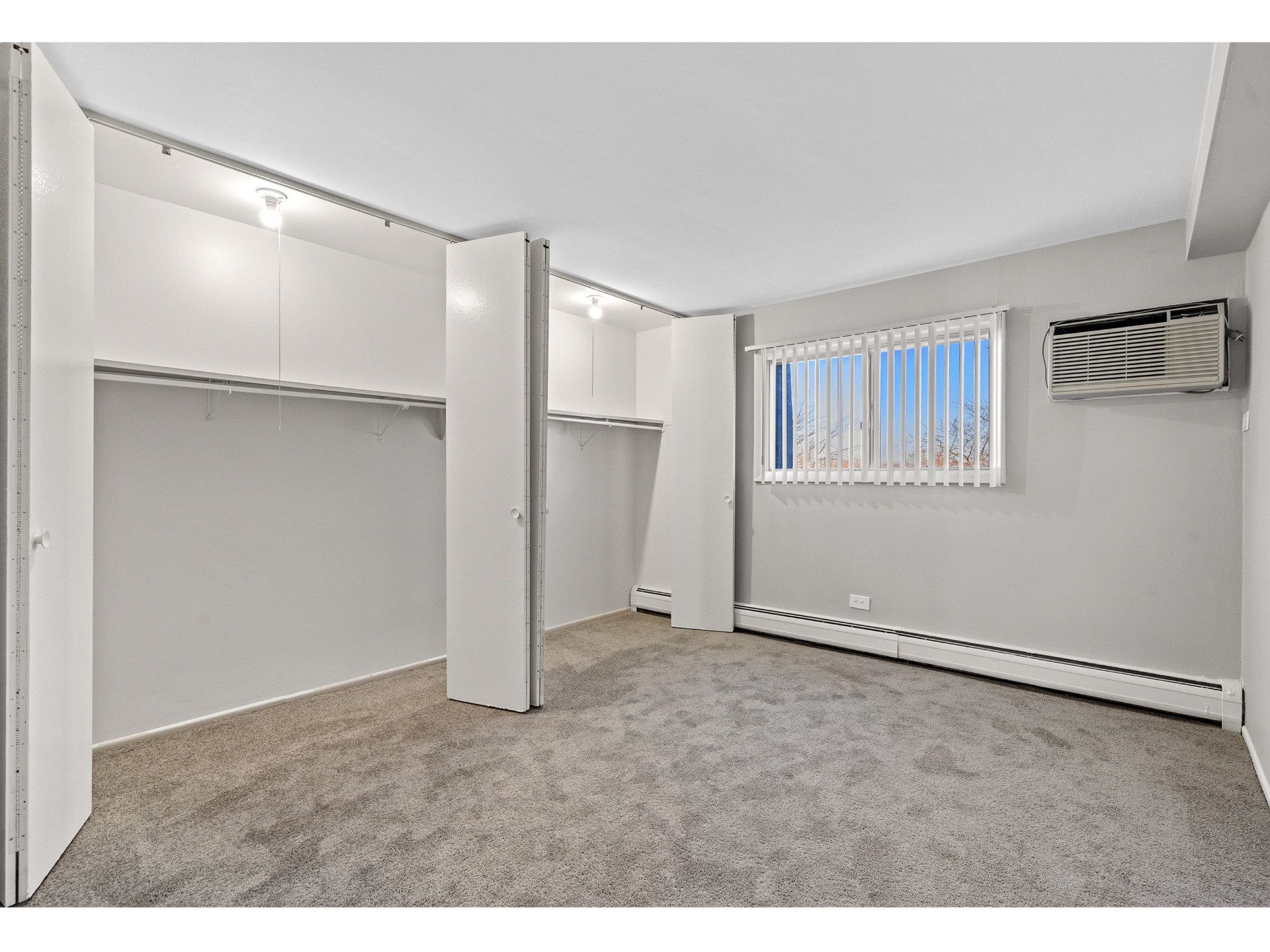 Empty bedroom with built-in closets, carpet, window with blinds, and a wall-mounted A/C unit.