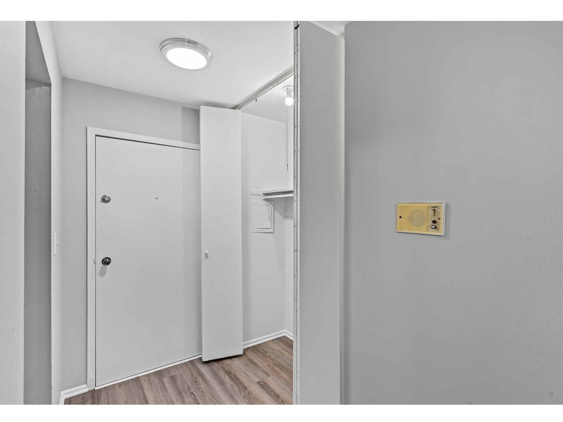 Entryway of an apartment with a white door, open closet, intercom panel, and wood floor.
