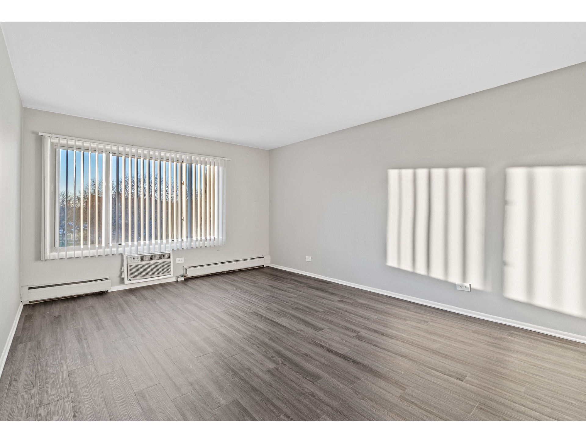 Empty living room with a large window and vertical blinds, gray walls, and wood-look flooring.