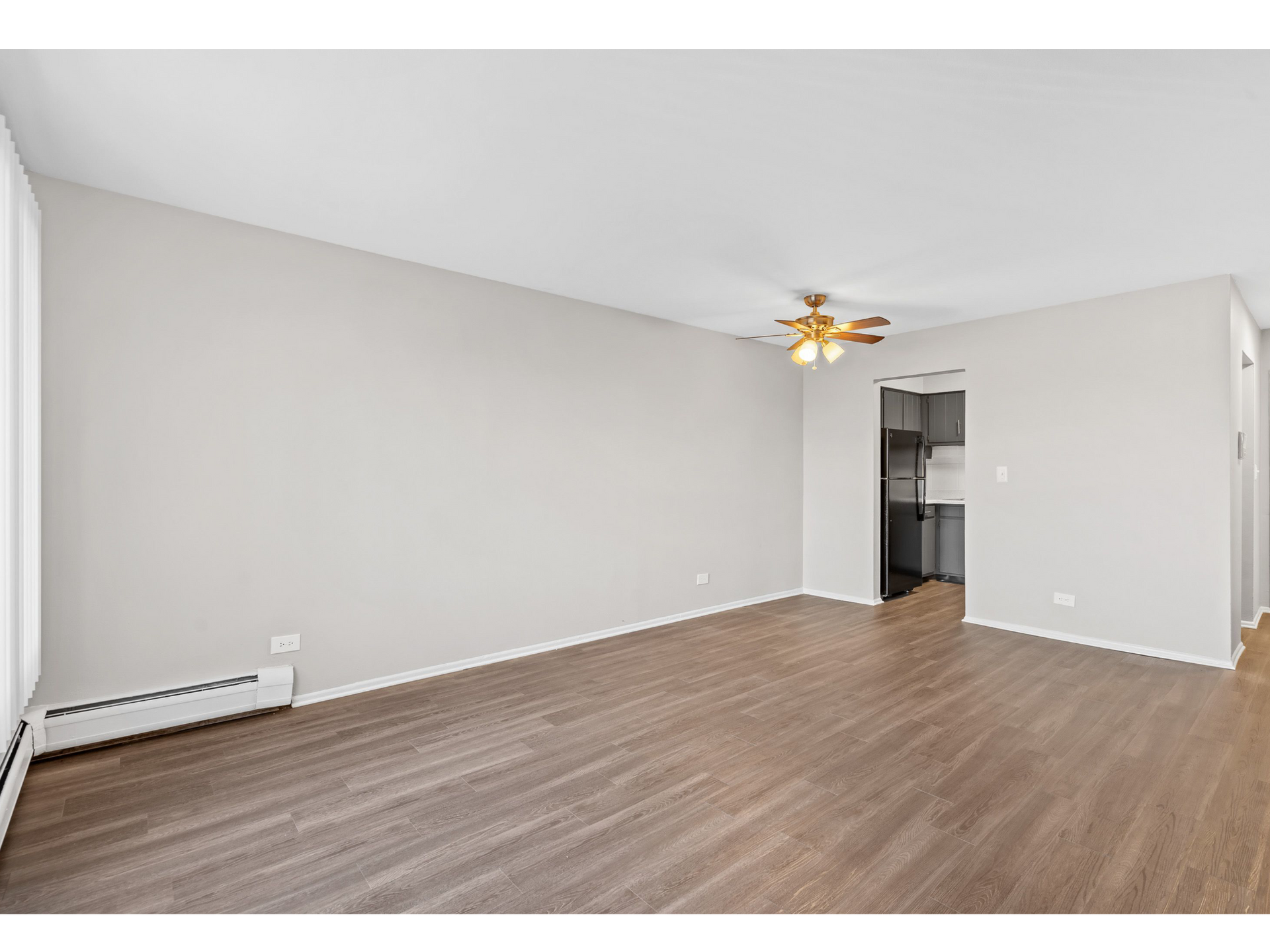 Empty living room with wood-look flooring, ceiling fan, and visible kitchen.