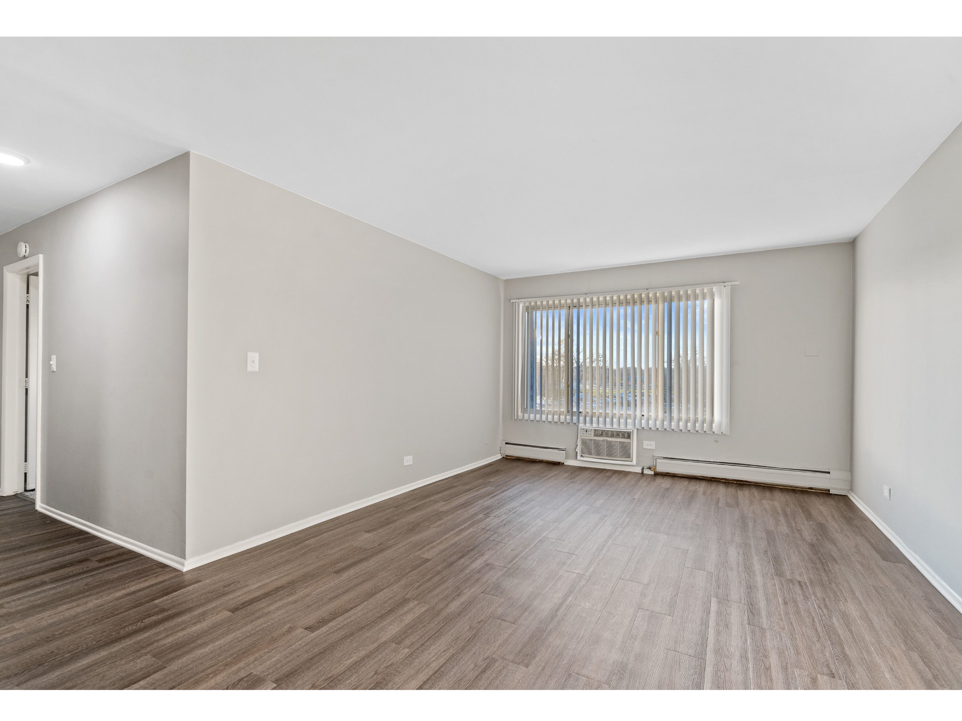 Bright living room with gray walls, wood-look flooring, and a large window with vertical blinds.