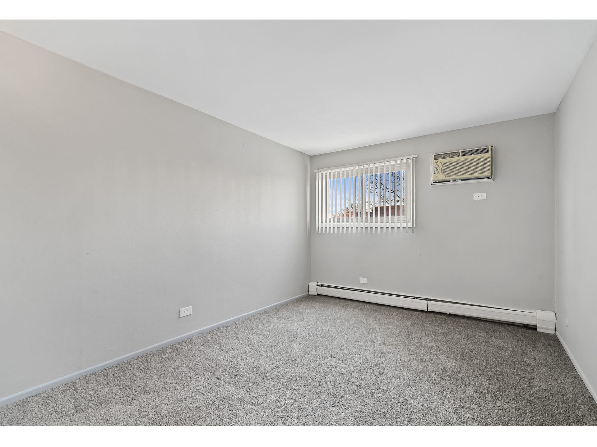 Small empty gray apartment room with a window, vertical blinds, and a wall-mounted air conditioner.