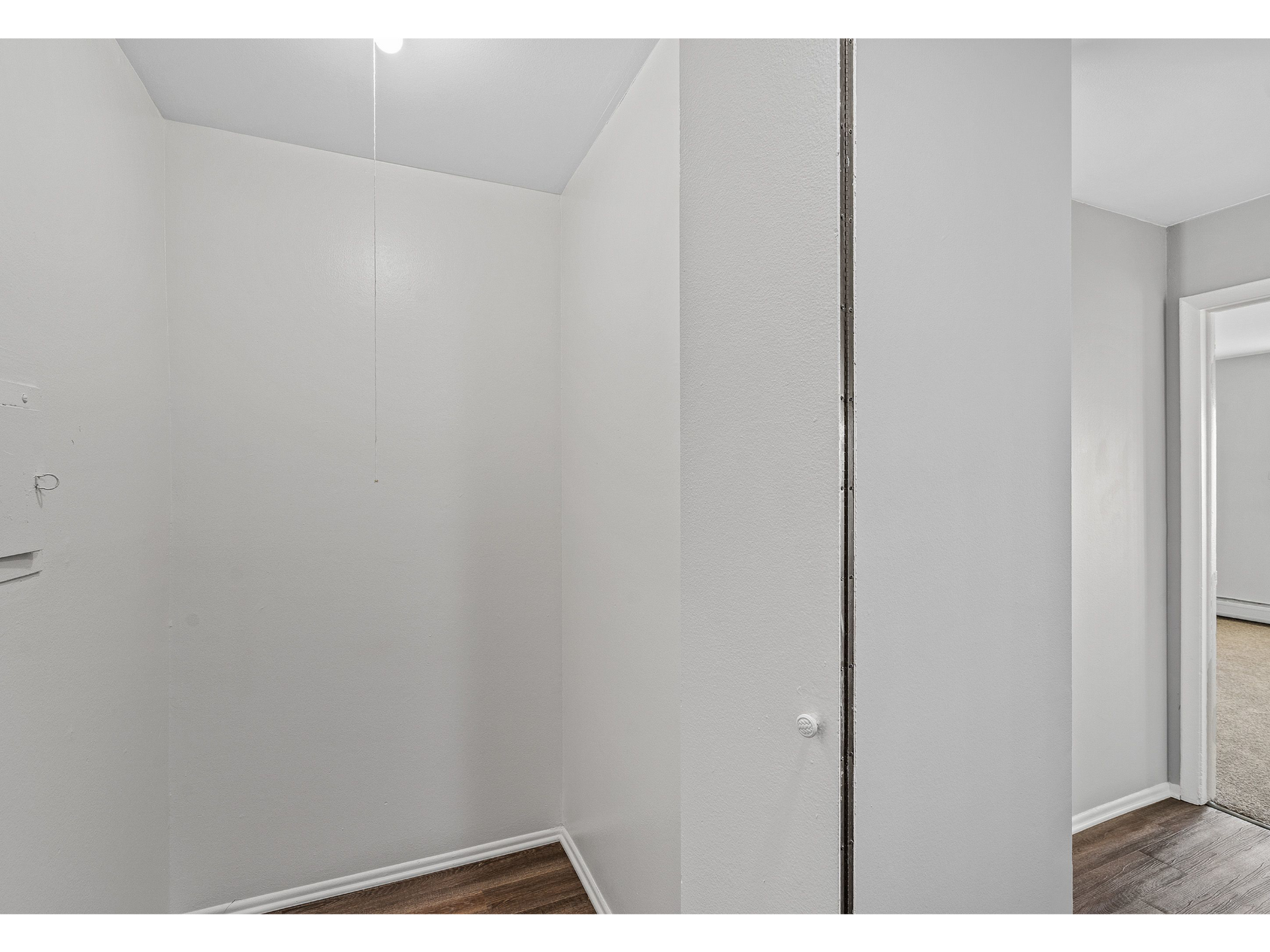 Empty white closet nook with pull-chain light and wood floor, opening to a carpeted room.