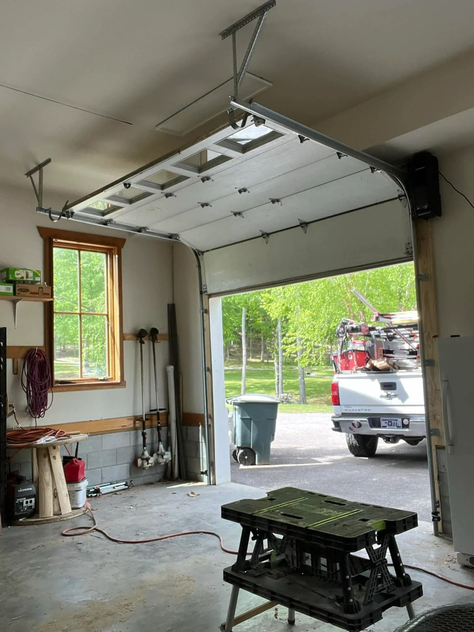 Garage door opening, revealing a truck and backyard. White door, grey concrete floor, work table in front.