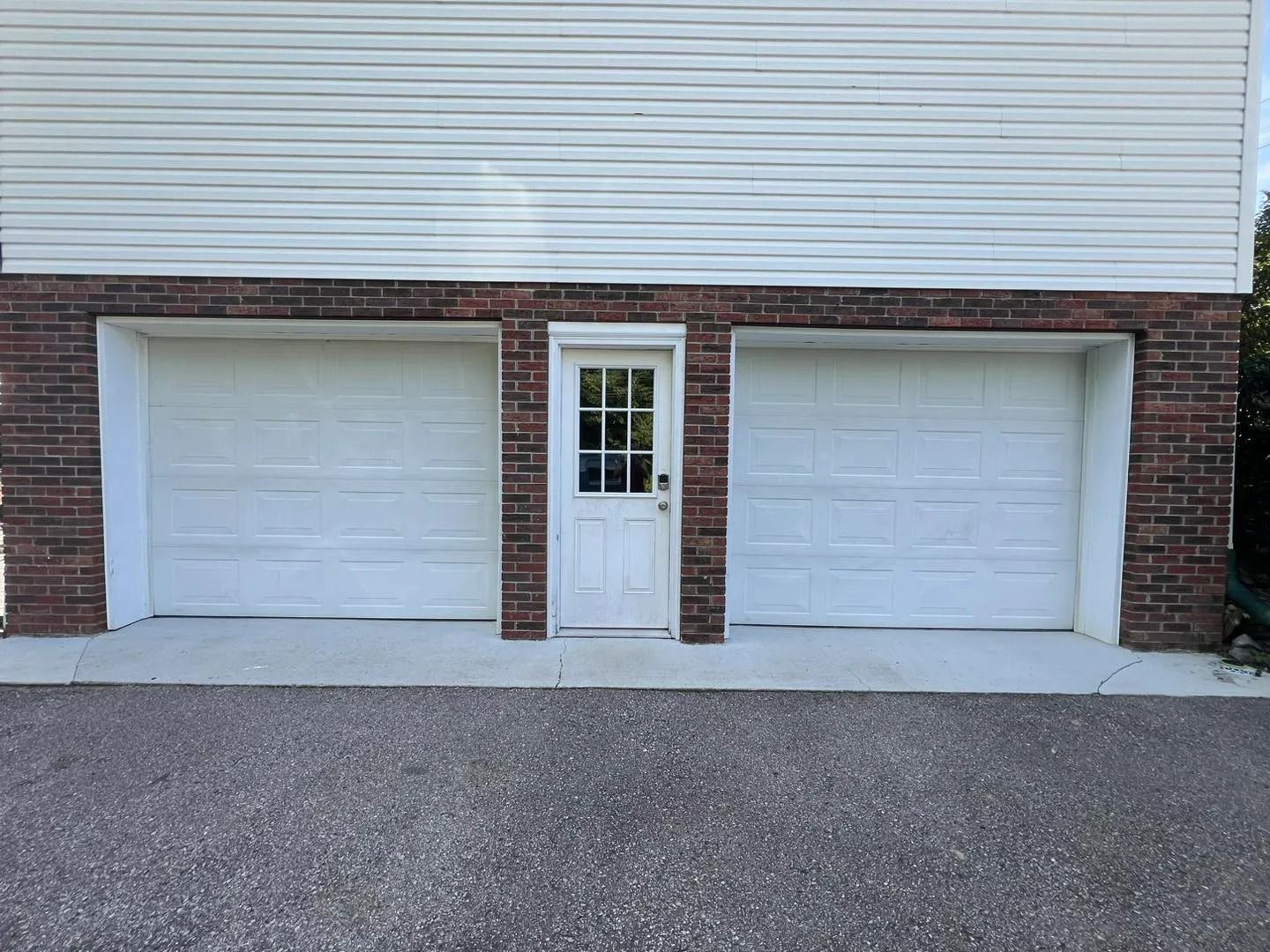 Two white garage doors and a white door with glass panels, set in brick, below a white-sided building.