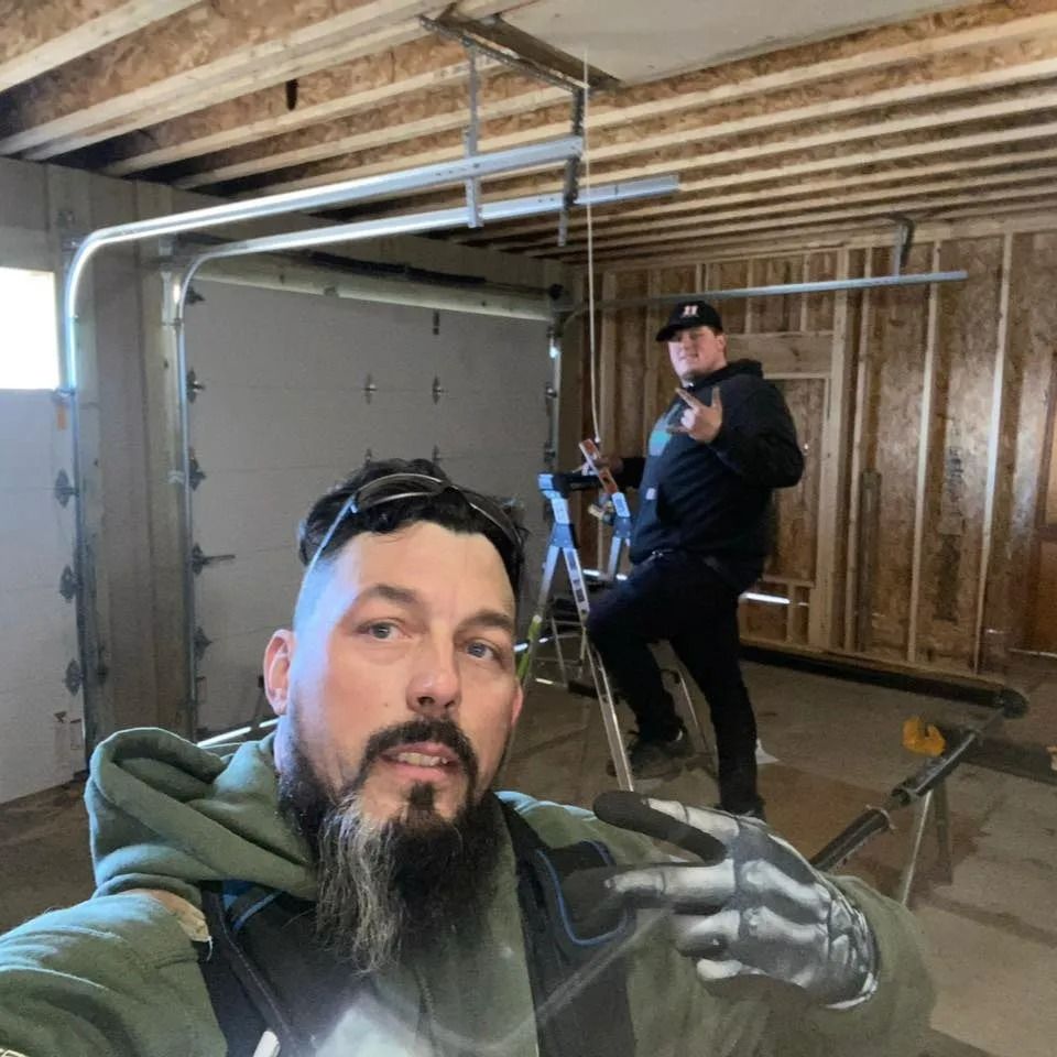 Two men in a garage installing a door. One takes a selfie, the other gestures.