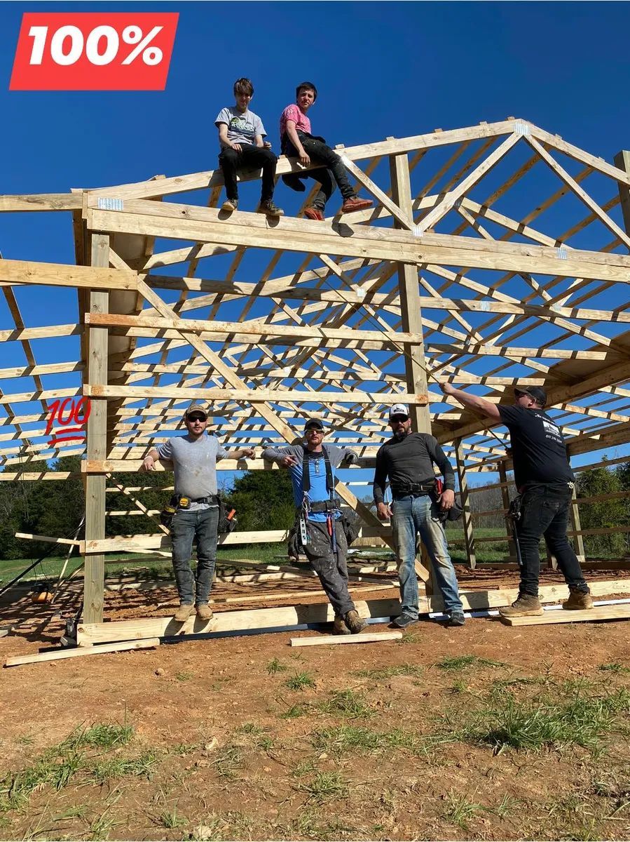 Five construction workers pose at a partially built barn on a sunny day; two sit on the roof.