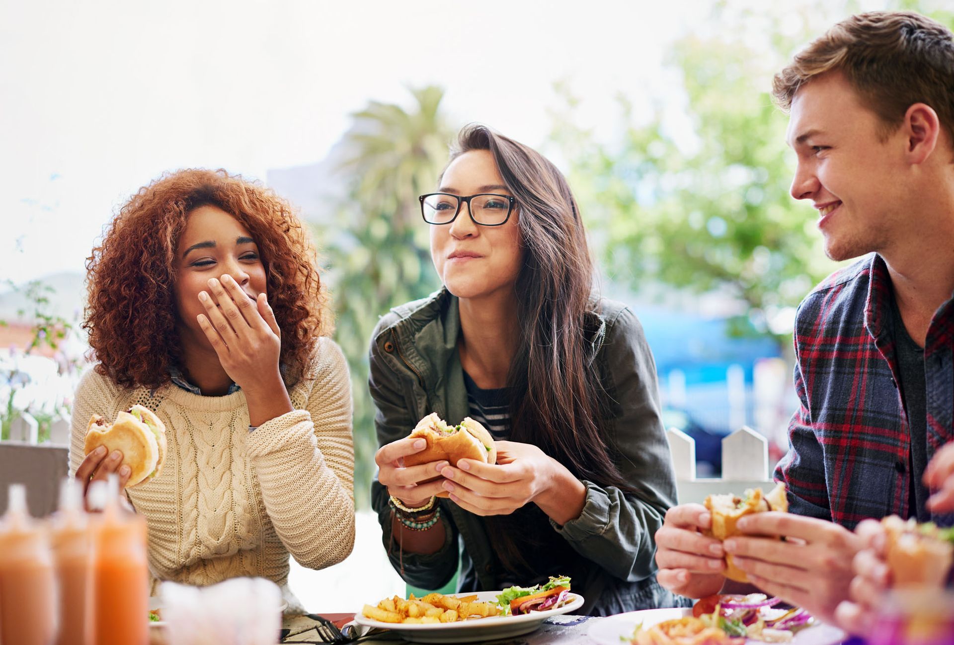 Three friends at an outdoor table eating burgers and laughing in a bright, casual setting