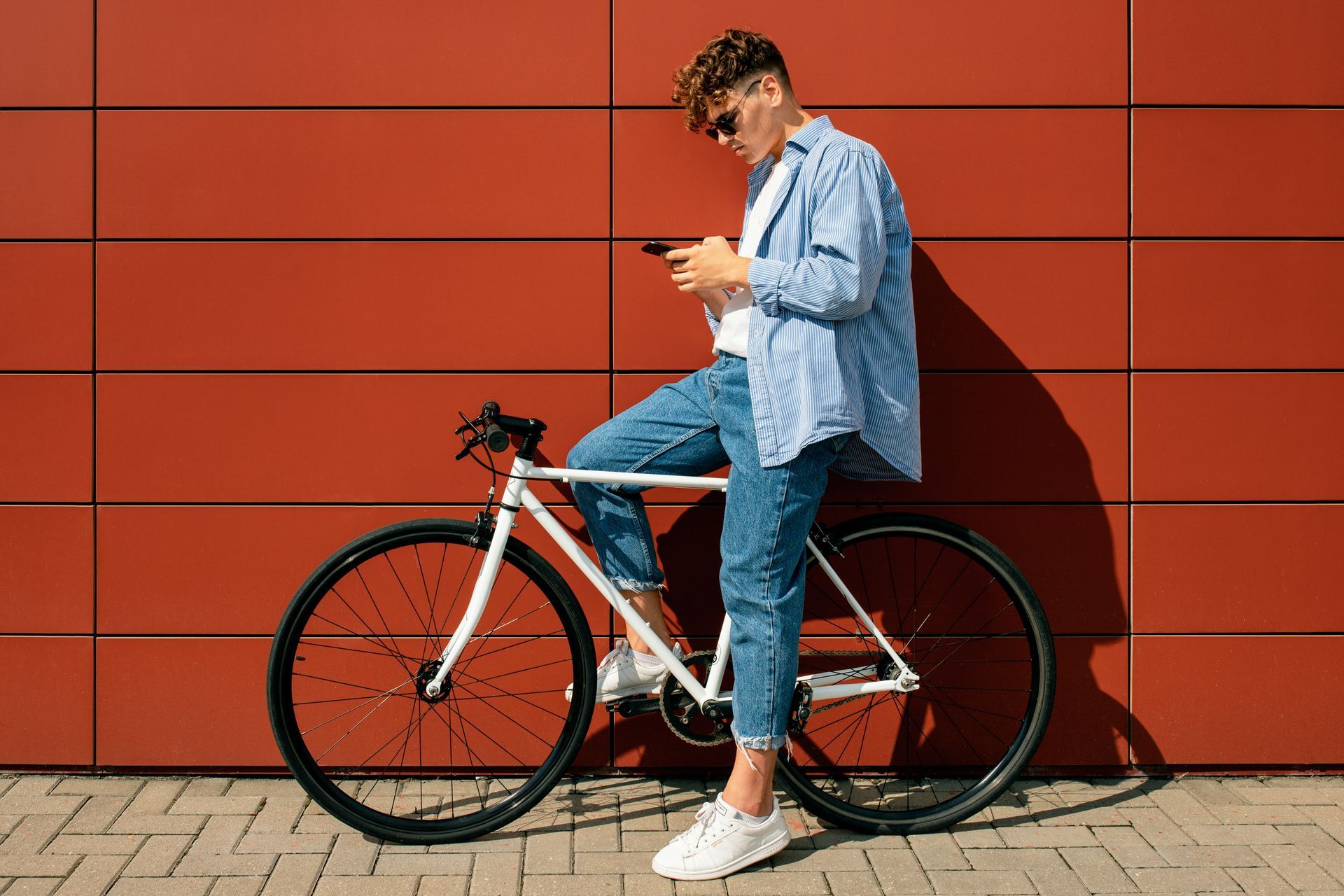 Person in blue shirt leaning on a bicycle against a red wall, looking at a phone